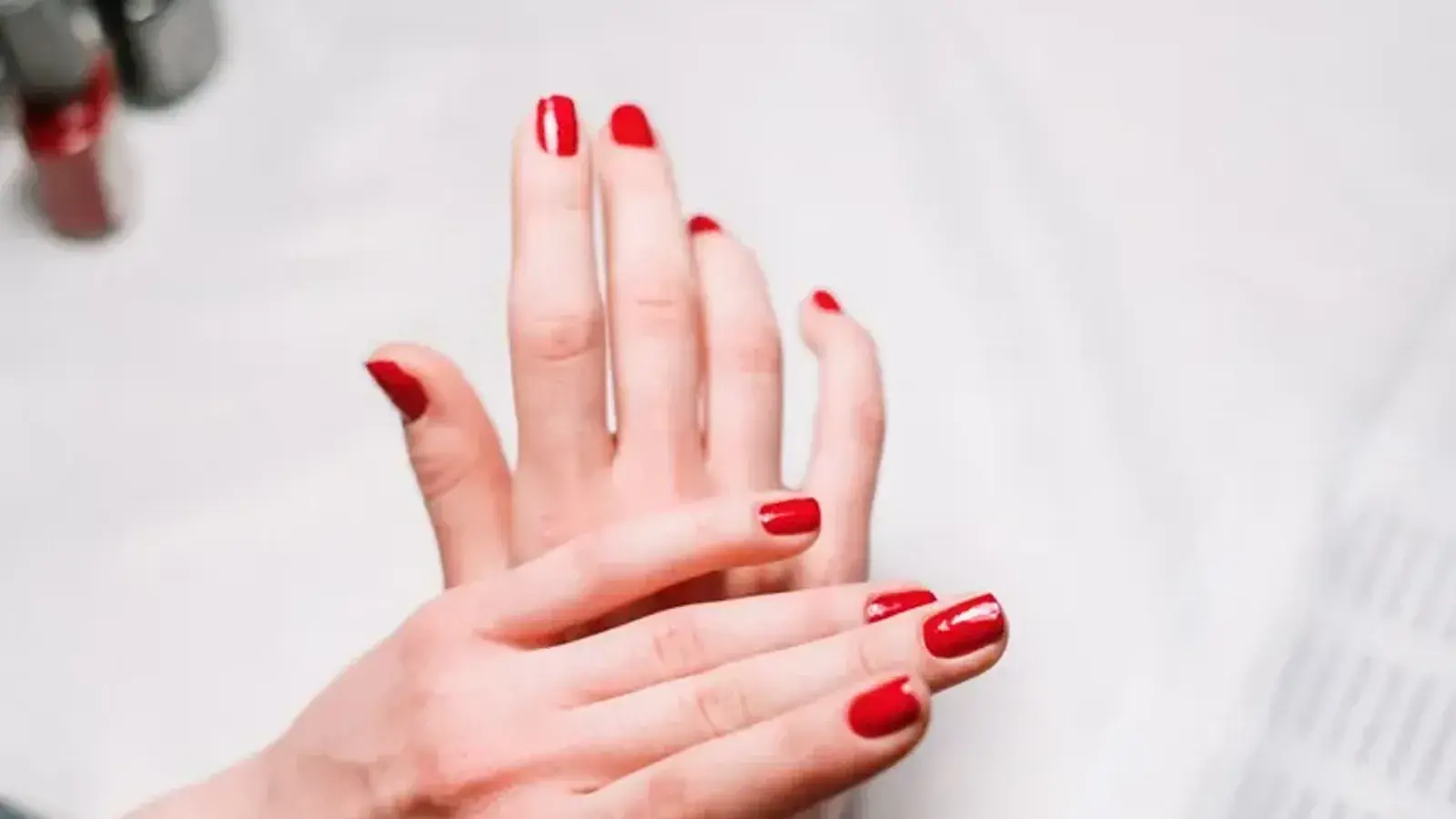 Two hands with glossy red nail polish resting on a white towel, with polish bottles in the background