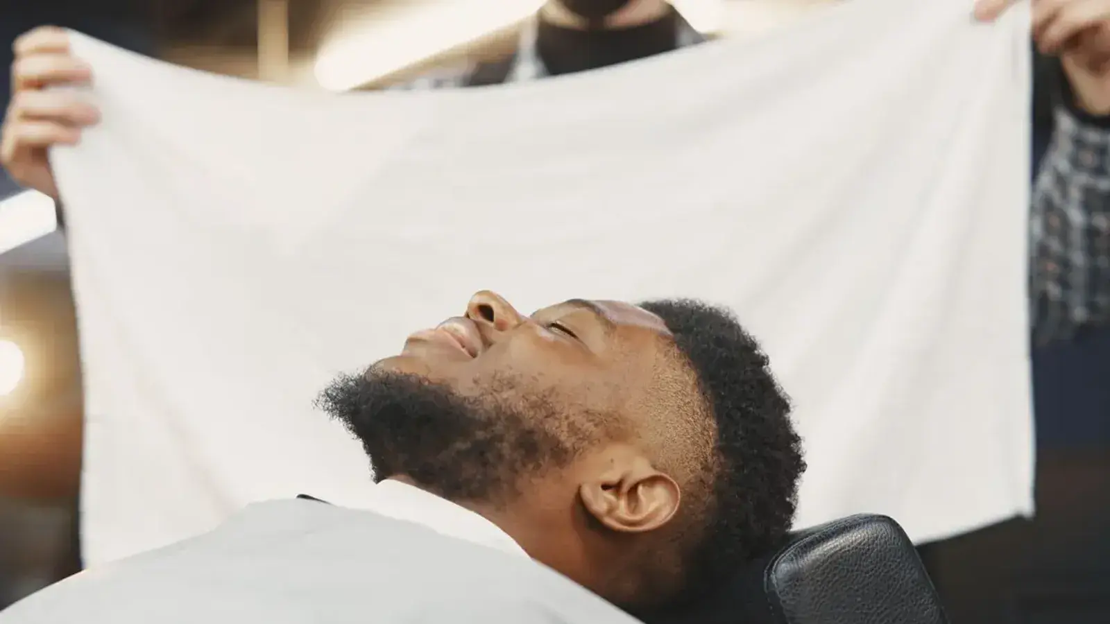 A smiling bearded man reclining in a barber chair as a towel is held up above his face