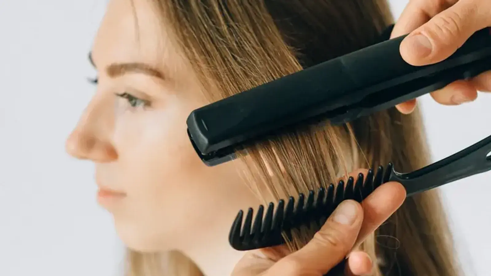 A stylist smoothing a section of a woman's long hair with a flat iron and a comb