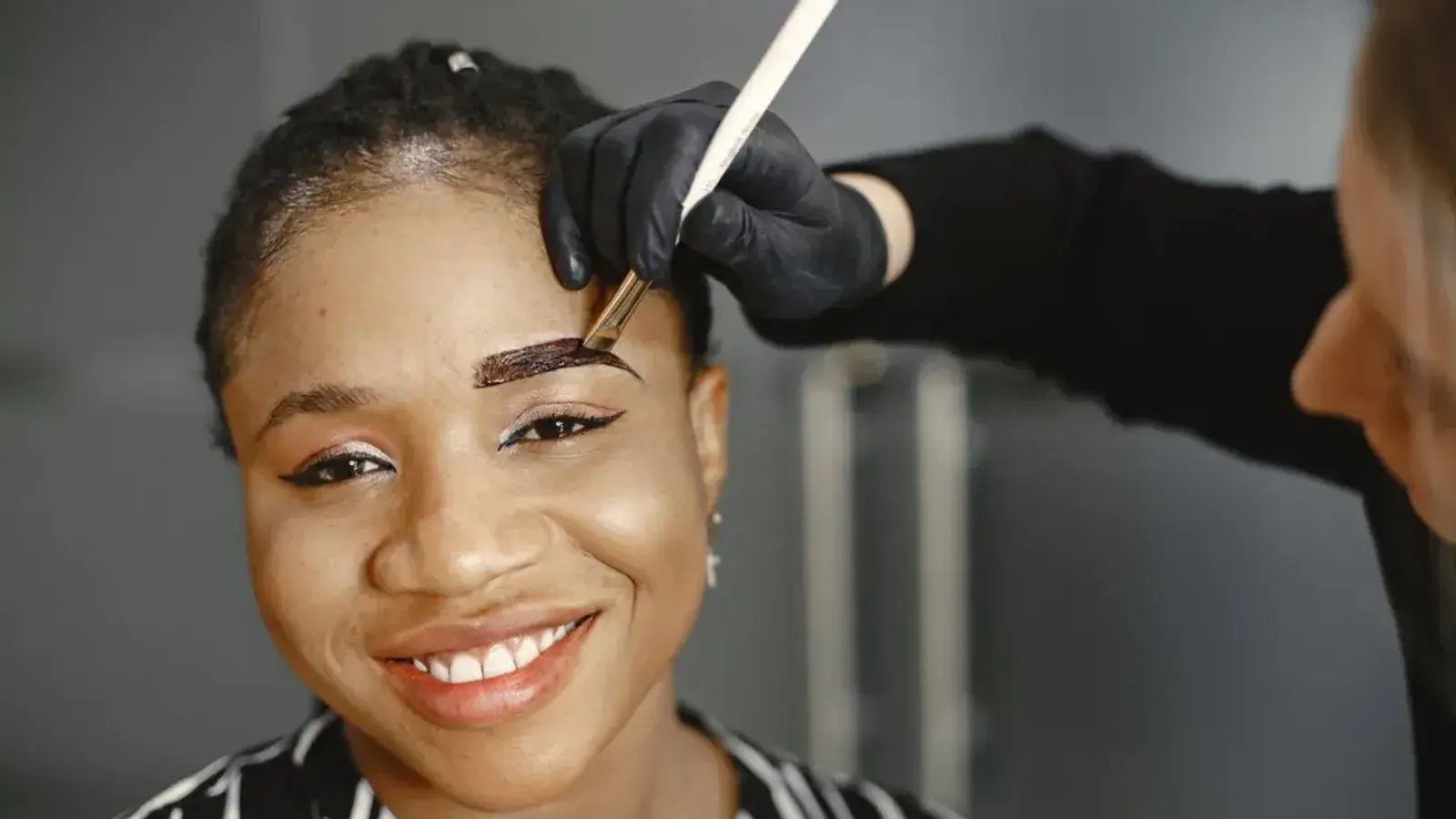 A beauty artist in black gloves brushing dark color onto a smiling woman's eyebrow