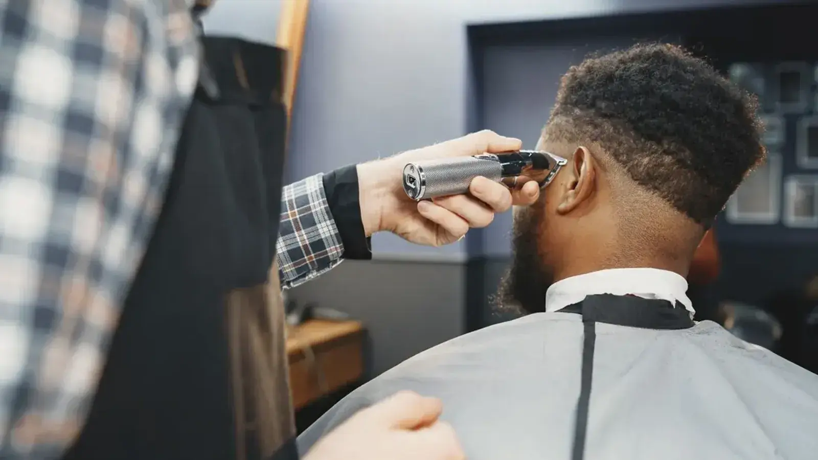 A barber in a plaid shirt trimming the side of a bearded man's hair with clippers