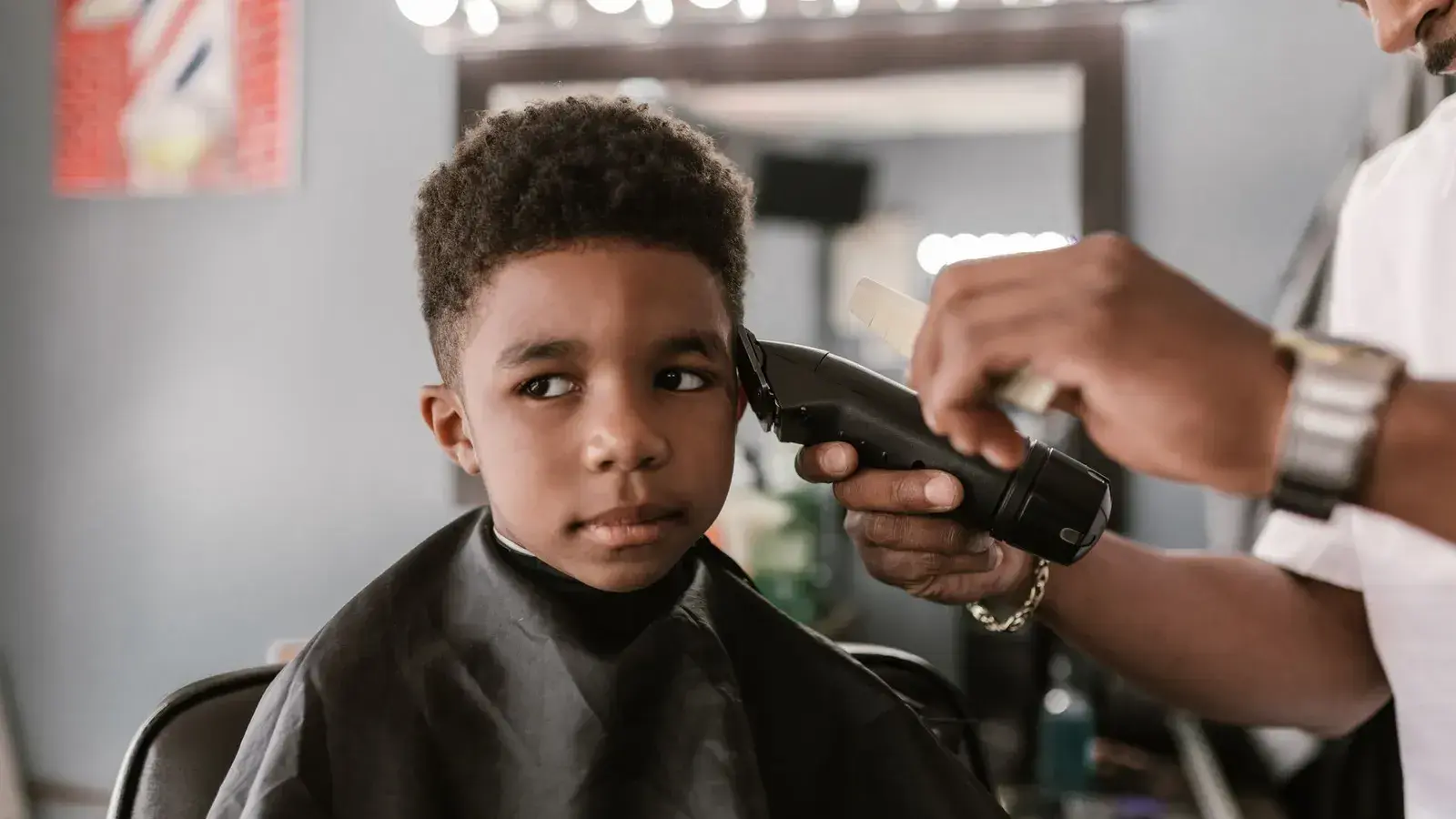 A barber trimming the side of a young boy's hair with clippers as the boy glances over