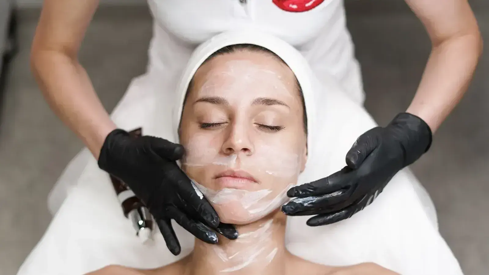 A woman lying back with her eyes closed while gloved hands spread a white cream across her face
