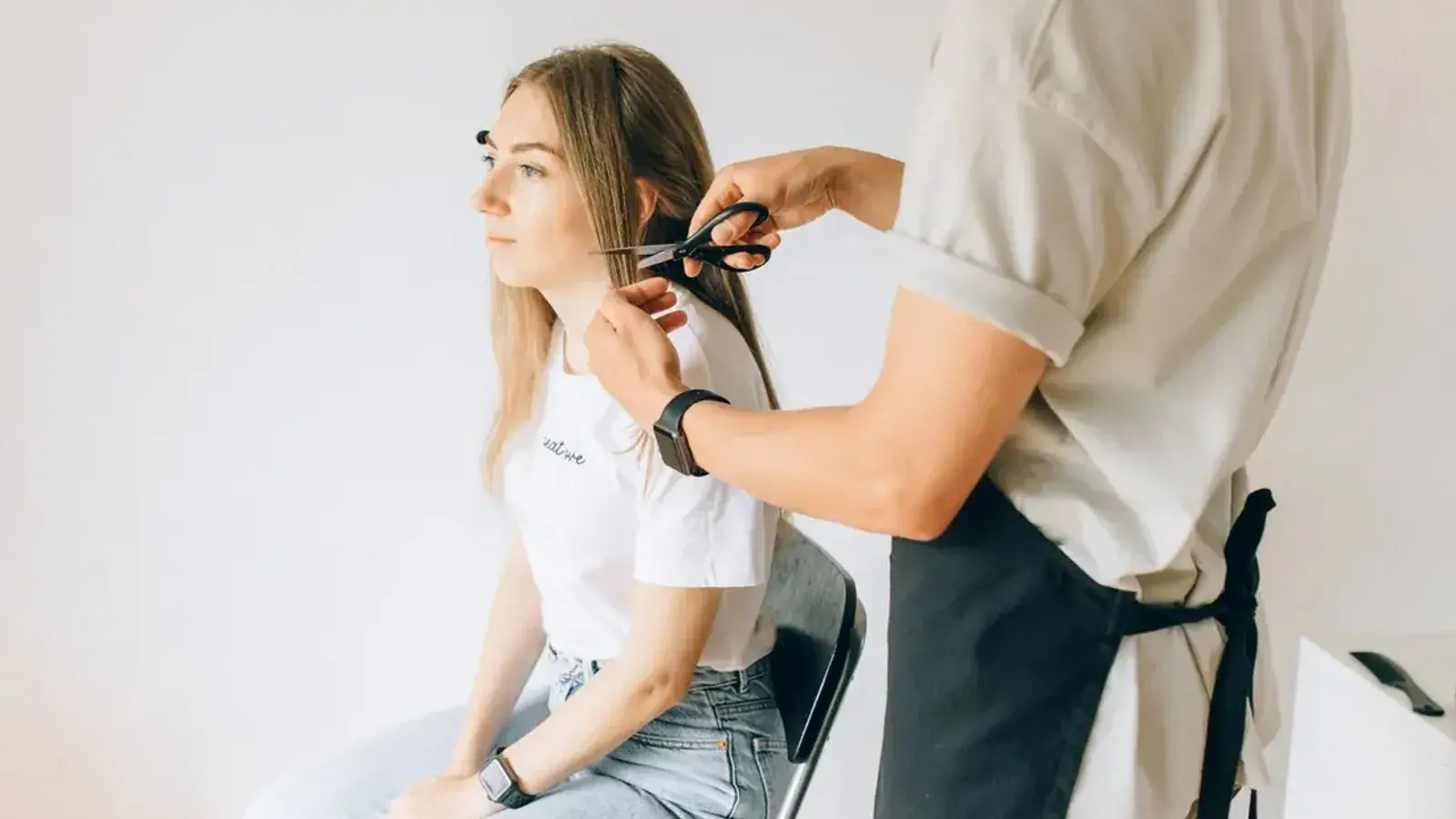 A stylist holding scissors to a section of a seated woman's long, light brown hair